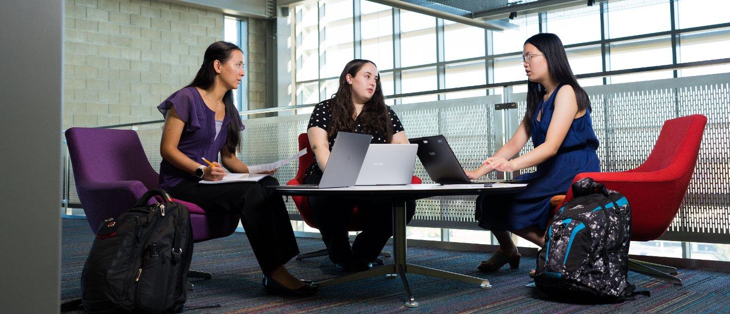 Three women seated around a table with open laptops in a lounge area.